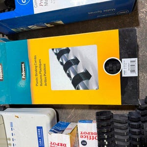 May include: A Fellowes brand box of black plastic binding combs with a yellow and teal design. The box features text reading Plastic Binding Combs, Reliures Plastiques, and Arillos Plasticos, alongside a graphic of the black binding spines.