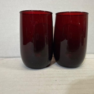 Könnte beinhalten: Zwei rubinrote Trinkgläser ohne Stiel. Die Gläser haben eine abgerundete, zylindrische Form mit glatter, glänzender Oberfläche. Die Farbe ist ein tiefes, sattes Rot, mit einem leichten Farbverlauf zu einem dunkleren Farbton am Boden. Sie sind leer und scheinen aus Glas zu sein.