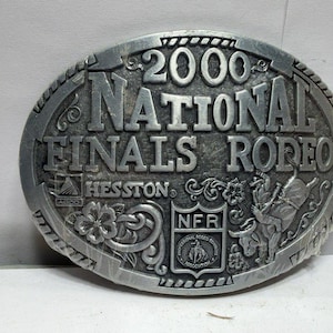 Op de afbeelding: Een ovale zilverkleurige metalen riemgesp met de in reliëf aangebrachte tekst 2000 NATIONAL FINALS RODEO. Het ontwerp bevat het HESSTON-logo, een NFR-schildembleem, bloemmotieven en een afbeelding van een rijder op een stier.