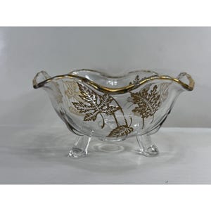 Op de afbeelding: Een heldere glazen kom met een gouden rand en drie kleine voetjes. De kom heeft een gouden bladerpatroon aan de zijkanten. De kom heeft een geschulpte rand.