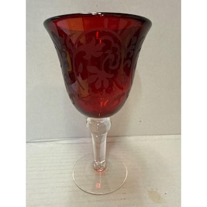May include: A ruby red wine glass with a clear stem and base. The bowl of the glass features an etched floral design. The glass is made of a transparent material, allowing light to pass through. The glass is designed for serving wine.