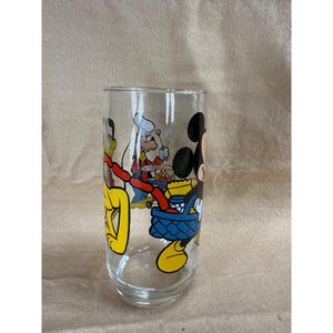 May include: A clear glass featuring a colorful cartoon illustration of Disney characters, including Mickey Mouse, Donald Duck, and Goofy. The characters are depicted in a picnic scene. The glass has a rounded shape and the words "Disney Productions" are printed on the bottom.