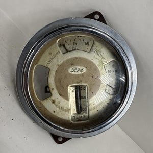 May include: Vintage Ford car dashboard gauge with a chrome bezel. The gauge features an oil pressure indicator, a temperature gauge, and a fuel gauge. The Ford logo is visible in the center. The gauge is circular and has a cream-colored face.