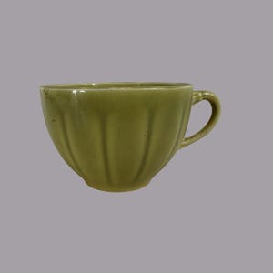 May include: A green ceramic teacup with a handle. The cup has a ribbed design on the exterior and a glossy finish. The teacup is set against a solid gray background.