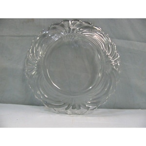 May include: A clear glass plate with a decorative, scalloped edge. The plate features a swirling, textured design around the rim, adding visual interest. The plate is circular and appears to be made of transparent glass.