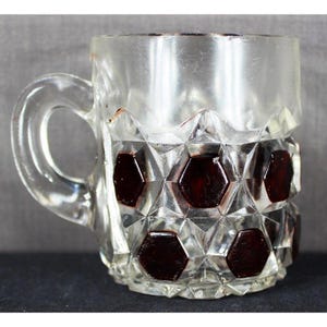 Könnte beinhalten: Transparenter Glasbecher mit Henkel. Der untere Teil hat ein geometrisches Design mit dunkelroten, sechseckigen Akzenten. Der obere Teil des Bechers ist glatt, und der Griff ist transparent. Für Getränke.