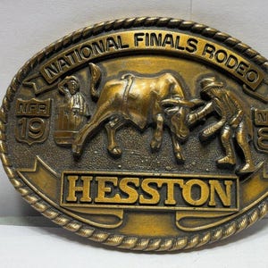 Op de afbeelding: Een ovale riemgesp in messingkleur met een reliëf van een stier en een rodeodeelnemer. De tekst luidt NATIONAL FINALS RODEO, HESSTON en NFR 1981. De rand heeft een touwachtige textuur en de achtergrond heeft een donkere, verweerde afwerking.