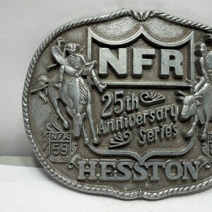 Op de afbeelding: Een ovale zilverkleurige metalen riemgesp met een reliëfontwerp van twee rodeoruiters op bokkende dieren. De centrale tekst luidt NFR 25th Anniversary Series en HESSTON, met de jaren 59 en 83 weergegeven in kleine schilden.