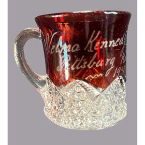 May include: A small, ruby-red glass mug with a clear handle and a diamond-cut base. The mug features etched white lettering that reads "Velma Kennedy Pittsburg, R. 1997". The top portion of the mug is a deep red color.