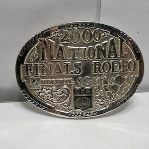 Peut inclure: Une boucle de ceinture ovale en métal argenté portant le texte en relief 2000 NATIONAL FINALS RODEO. Le design comprend le logo Hesston, un emblème en forme de bouclier NFR et des motifs complexes de volutes florales sur les bords.