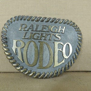 May include: Oval-shaped belt buckle with the words "RALEIGH LIGHTS RODEO" in raised gold lettering. The buckle has a textured gray background and a twisted rope border. A vintage western-themed accessory.