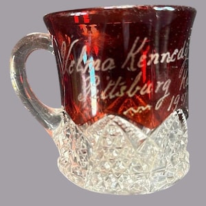 May include: A small, ruby-red glass mug with a clear handle and a decorative, cut-glass base. The mug features etched white lettering that reads "Velma Kennedy Pittsburg, 1997". The glass has a vintage aesthetic.