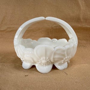 Puede incluir: Una cesta de vidrio lechoso blanco con asa. La cesta tiene forma de flor con un borde festoneado y descansa sobre tres pequeños pies. El asa se arquea elegantemente sobre la cesta. La cesta es probablemente vintage.