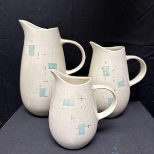 Peut inclure: Trois pichets en céramique mouchetée de tailles différentes. Les pichets sont blanc cassé avec un motif de rectangles bleus et de signes plus rouges. Chaque pichet a une anse et un bec verseur.