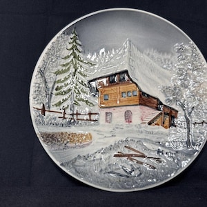 May include: A decorative ceramic plate featuring a winter scene. The plate depicts a snow-covered cabin with a brown wooden exterior, surrounded by trees and a fence. The color palette includes shades of gray, brown, and white, creating a serene, wintry atmosphere.