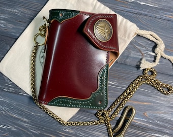 Pode incluir: Uma carteira de couro bordô com acabamentos verdes e uma corrente de latão. A carteira apresenta um emblema decorativo de latão e é exibida em uma superfície de madeira. Uma bolsa branca com cordão também está presente.