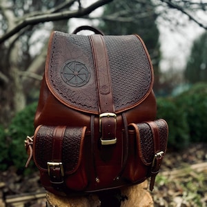 Könnte beinhalten: Ein brauner Lederrucksack mit einer großen Fronttasche und zwei kleineren Seitentaschen. Der Rucksack hat ein dekoratives geprägtes Design auf der Vorderseite, einschließlich eines kreisförmigen Symbols mit einem radförmigen Muster. Der Rucksack steht auf einem Baumstumpf.