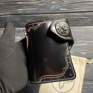May include: Black leather wallet with a silver-colored metal emblem featuring a Native American chief skull design. The wallet has white stitching and a light brown trim. A small, off-white drawstring bag is in the background.