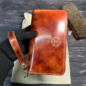 Könnte beinhalten: Eine braune Ledergeldbörse mit Handschlaufe und goldenem Reißverschluss. Die Geldbörse hat eine strukturierte Oberfläche und ein goldenes Emblem. Die Handschlaufe ist mit einem goldenen Verschluss befestigt. Die Geldbörse wird von einer behandschuhten Hand gehalten.