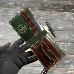 Può includere: Un portafoglio in pelle a due ante nei toni del verde e del marrone. Il portafoglio presenta un design cucito, più fessure per carte e una piccola tasca per monete. Un emblema dorato è visibile sulla sezione verde. Il portafoglio è tenuto da una mano guantata.