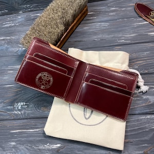 Può includere: Portafoglio in pelle bordeaux aperto, con scomparti per carte, appoggiato su un sacchetto con coulisse color crema. Visibili anche una spazzola con manico in legno e un accessorio in pelle. Il portafoglio presenta un logo dorato in rilievo.