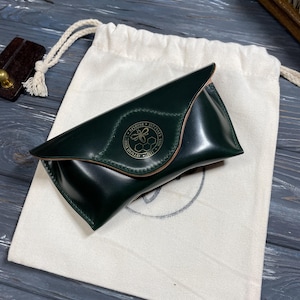 Könnte beinhalten: Ein dunkelgrünes Lederetui für Brillen mit goldenem Emblem, auf einem weißen Kordelzugbeutel. Das Etui ist geschlossen, hat eine geschwungene Form und eine genähte Kante. Der Beutel liegt auf einer dunklen Holzoberfläche.