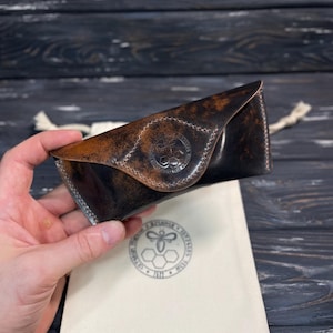 Puede incluir: Un estuche para gafas de cuero marrón oscuro con aspecto envejecido y cierre de solapa triangular. La solapa frontal presenta un logotipo circular en relieve con una abeja y un patrón de panal. Incluye una bolsa de tela de color crema.