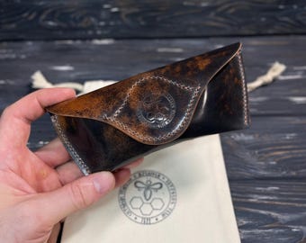 May include: A dark brown, distressed leather glasses case with a triangular flap closure. The case features a circular embossed logo of a bee and honeycomb pattern on the front flap. It is accompanied by a cream-colored fabric drawstring pouch.