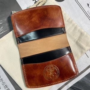 Può includere: Un portafoglio in pelle marrone con dettagli neri e un'emblema dorato. Il portafoglio ha più fessure per carte e un vano centrale. La pelle ha una finitura invecchiata e le cuciture sono visibili sui bordi. Il portafoglio è aperto, rivelando l'interno.