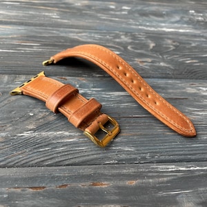 Può includere: Un cinturino da orologio in pelle marrone con una fibbia dorata. Il cinturino è progettato per un Apple Watch.