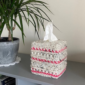 Pode incluir: Uma capa de caixa de lenços de tecido crochet com listras rosa e brancas. A capa é feita com um fio texturizado e tem uma forma quadrada.