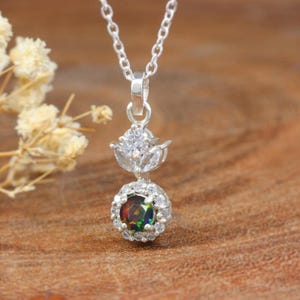 May include: A silver pendant necklace featuring a round, iridescent gemstone surrounded by small, clear stones. The pendant has a floral design with a clear, round stone at the top. The necklace is on a silver chain.