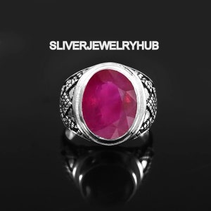 Könnte beinhalten: Ein silberner Ring mit einem ovalen Rubin-Edelstein. Der Ring hat ein detailliertes Design mit komplizierten Mustern um den Stein herum.