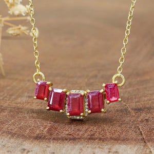 May include: A gold necklace with five rectangular, deep red gemstones. The central gemstone is framed by small, clear stones. The necklace is suspended from a fine gold chain, presenting a classic and elegant design.