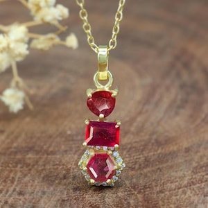 Könnte beinhalten: Eine goldene Kettenkette mit einem Anhänger mit drei roten Edelsteinen. Der Anhänger besteht aus einem herzförmigen Stein, einem quadratischen Stein und einem sechseckigen Stein.