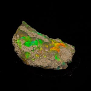 Könnte beinhalten: Ein roher, natürlicher Opal mit grünem, orangem und irisierendem Schimmer. Der Opal ist in eine Matrix aus grauem Gestein eingebettet.