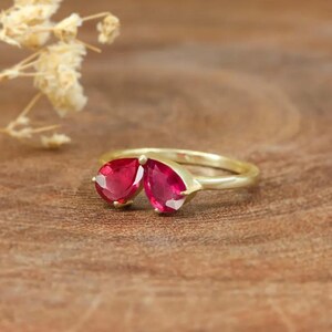 Könnte beinhalten: Ein goldener Ring mit zwei birnenförmigen Rubinen. Die Edelsteine sind so gefasst, dass sie sich zu berühren scheinen.