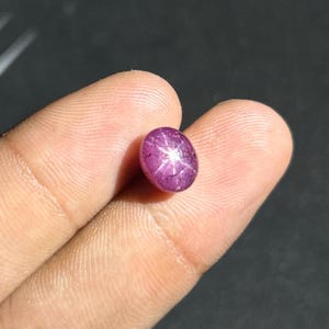 Könnte beinhalten: Ein polierter, ovalförmiger Sternrubin-Edelstein. Der Edelstein hat eine tiefrosa-rote Farbe mit einem sechsstrahligen Sternmuster auf der Oberfläche. Der Edelstein wird zwischen zwei Fingern vor einem dunklen Hintergrund gehalten.