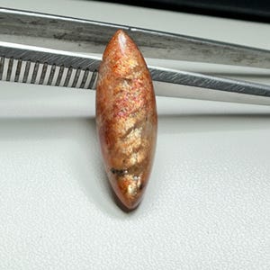 Könnte beinhalten: Ein polierter, oval geformter Edelstein mit einem warmen, orangebraunen Farbton und schimmernden Einschlüssen. Der Edelstein wird von einer silbernen Pinzette gehalten, die seine längliche Form und natürliche Schönheit zeigt. Die Oberfläche des Steins reflektiert das Licht.