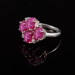 Könnte beinhalten: Ein silberner Ring mit fünf rosafarbenen sternförmigen Rubinen und kleinen Diamanten, die in die Ringschiene eingefasst sind.
