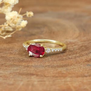 Könnte beinhalten: Ein goldener Ring mit einem roten ovalen Edelstein und kleineren weißen Edelsteinen, die in einer Bandfassung gefasst sind.