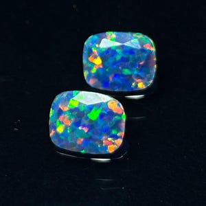 Könnte beinhalten: Zwei kissenförmige schwarze Opale mit lebendigen Farben. Die Opale zeigen eine Mischung aus Blau, Grün, Orange und Gelb vor dunklem Hintergrund. Die Edelsteine sind facettiert und fangen das Licht ein.