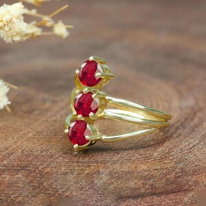 Könnte beinhalten: Ein goldener Ring mit drei roten Edelsteinen, die in einem versetzten Muster angeordnet sind. Der Ring besteht aus drei miteinander verbundenen Goldbändern.