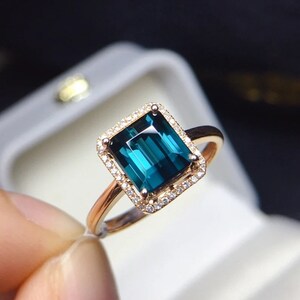 Könnte beinhalten: Ein goldener Ring mit einem großen smaragdgeschliffenen blauen Edelstein, der von einem Heiligenschein aus kleineren Diamanten umgeben ist.