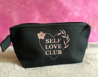 May include: Black canvas makeup bag with a zipper closure. The bag has a pink heart design with the words "SELF LOVE CLUB" inside the heart.