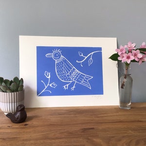 Op de afbeelding: Een witte linosnede van een gestileerde vogel op een tak met een blauwe achtergrond. De print is gesigneerd "K. James".