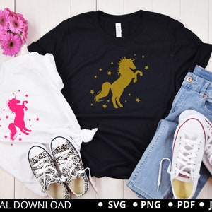 Puede incluir: Dos camisetas blancas con un dise&ntilde;o de unicornio. Una camiseta presenta un unicornio rosa con estrellas, la otra presenta un unicornio dorado con estrellas. Las camisetas est&aacute;n dispuestas sobre una superficie de madera blanca con un par de jeans y zapatillas blancas.