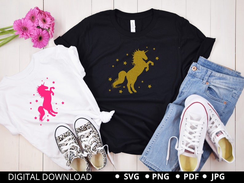 Puede incluir: Dos camisetas blancas con un dise&ntilde;o de unicornio. Una camiseta presenta un unicornio rosa con estrellas, la otra presenta un unicornio dorado con estrellas. Las camisetas est&aacute;n dispuestas sobre una superficie de madera blanca con un par de jeans y zapatillas blancas.