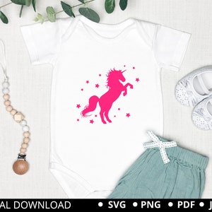 Puede incluir: Un body blanco con una silueta de unicornio rosa y estrellas. El unicornio se levanta sobre sus patas traseras con su cuerno apuntando hacia arriba. El body est&aacute; sobre un fondo blanco con un par de zapatos de beb&eacute; blancos y un mordedor de beb&eacute; de madera.