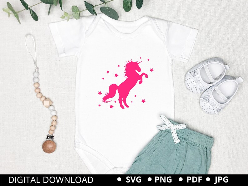 Puede incluir: Un body blanco con una silueta de unicornio rosa y estrellas. El unicornio se levanta sobre sus patas traseras con su cuerno apuntando hacia arriba. El body est&aacute; sobre un fondo blanco con un par de zapatos de beb&eacute; blancos y un mordedor de beb&eacute; de madera.